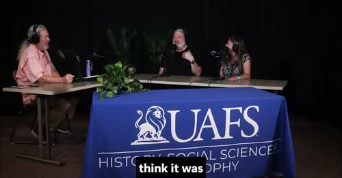 Three people participate in a recorded panel discussion at a table with a University of Arkansas–Fort Smith tablecloth. A host sits on the left, interviewing two guests seated on the right.
