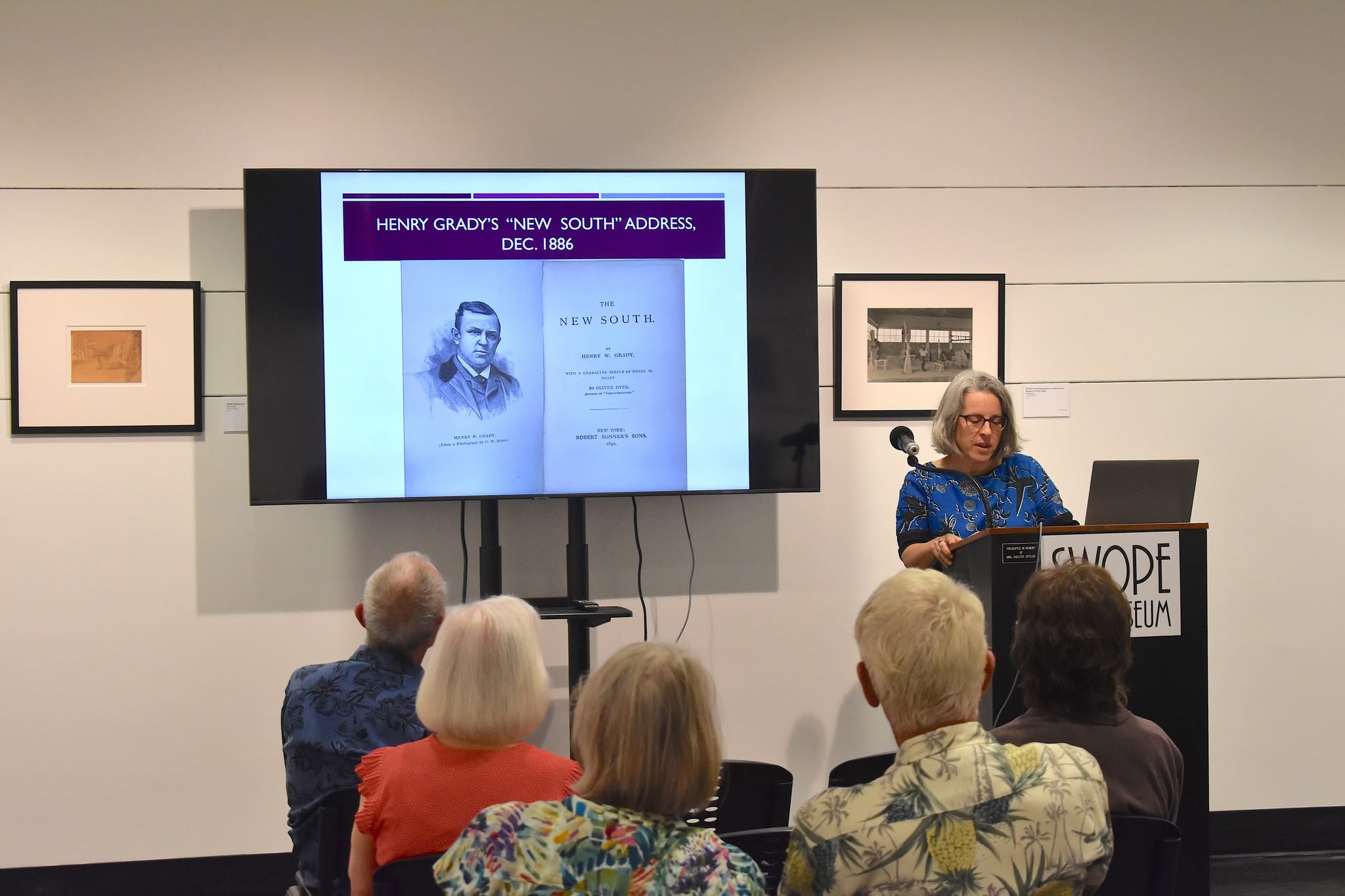 A speaker stands at a podium in a museum gallery, presenting to a seated audience. A large screen displays a slide titled "Henry Grady's 'New South' Address, Dec. 1886," featuring a portrait of Henry Grady and the title page of The New South.