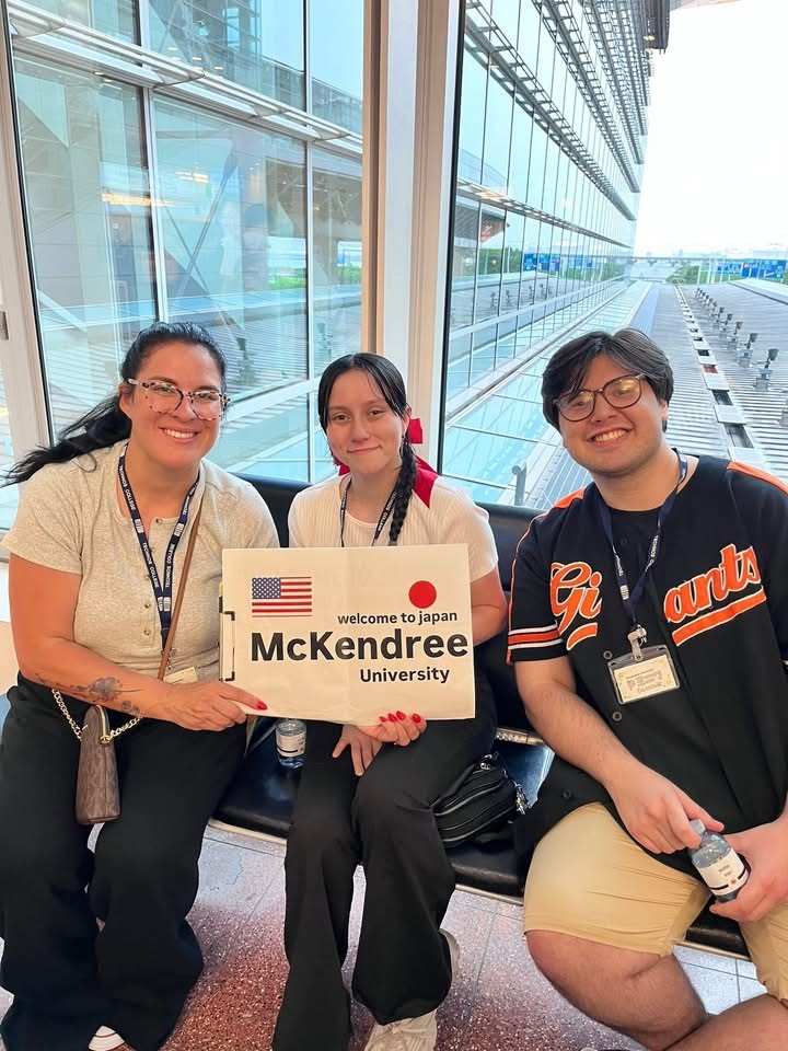 Three members of the McKendree University travel group sit in an airport terminal holding a “Welcome to Japan” sign